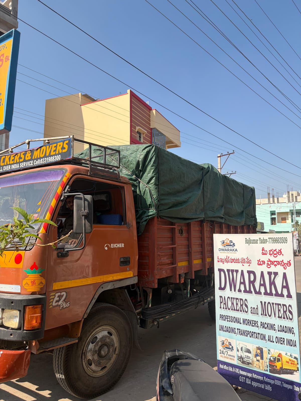 A loaded and tarpaulin-covered Dwaraka Packers and Movers truck parked beside the company's branded signboard