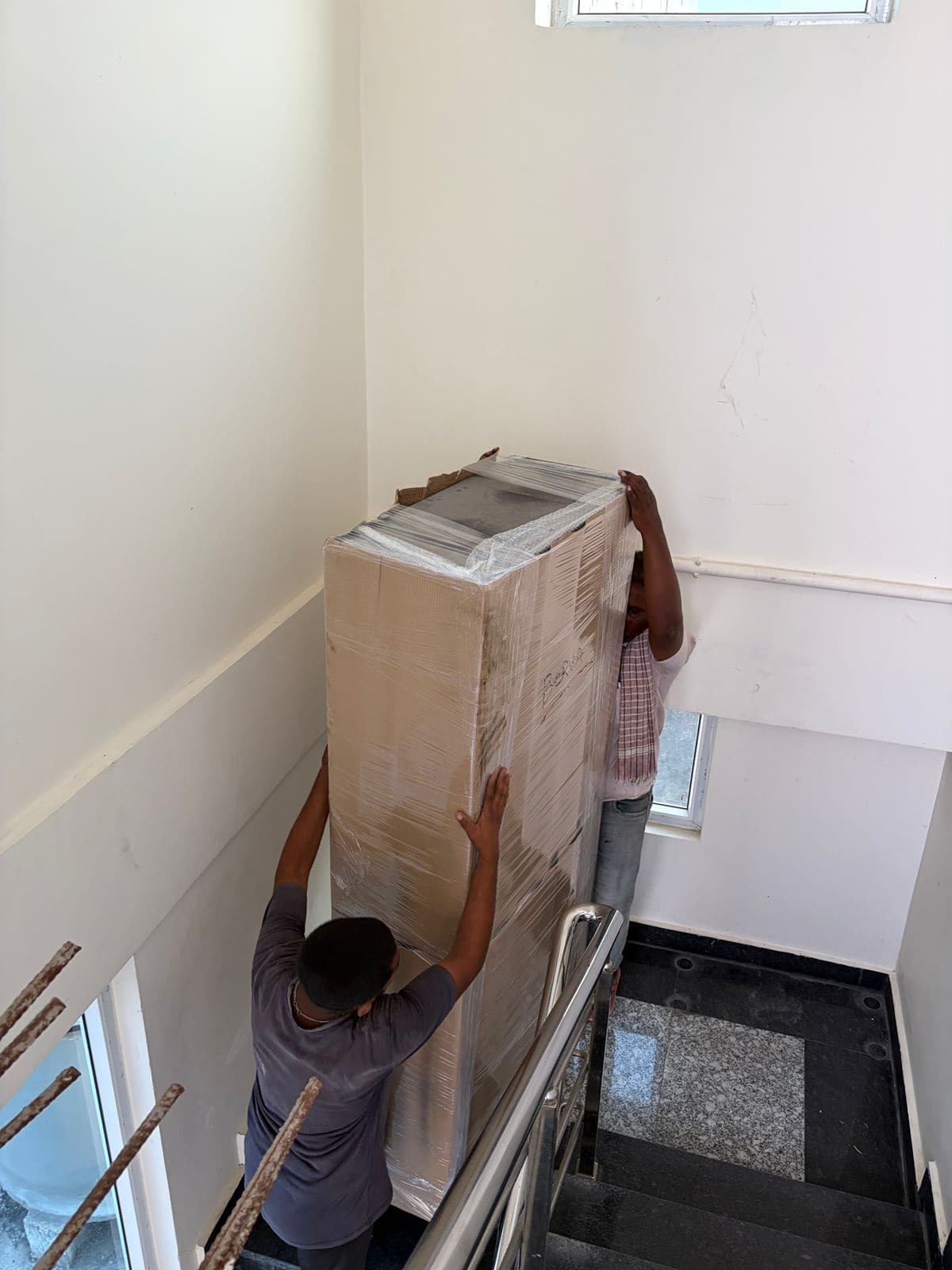 Two Dwaraka movers carefully carrying a wrapped refrigerator down an apartment staircase